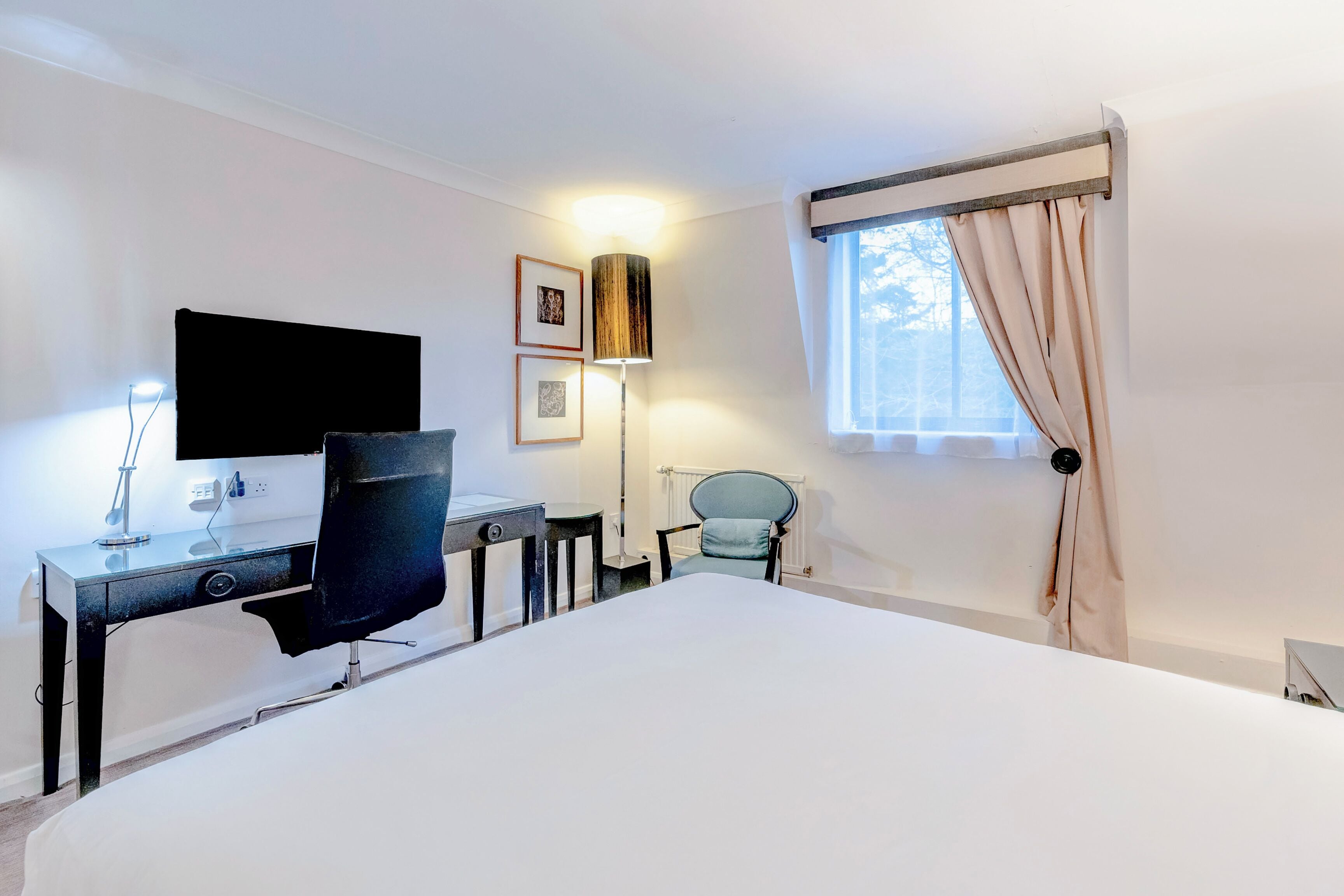 A hotel room with a bed, a desk with a chair, a lamp, and a window with curtains.