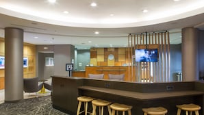 A modern reception area with a curved desk, wooden stools, a TV screen, and a seating area with a chair. - Springhill Suites by Marriott Billings (Billings)