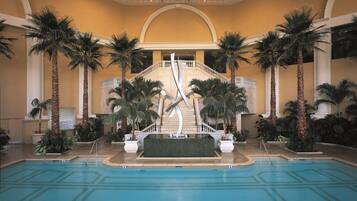 Una piscina interior con una escalera central, palmeras y una pared de azulejos.