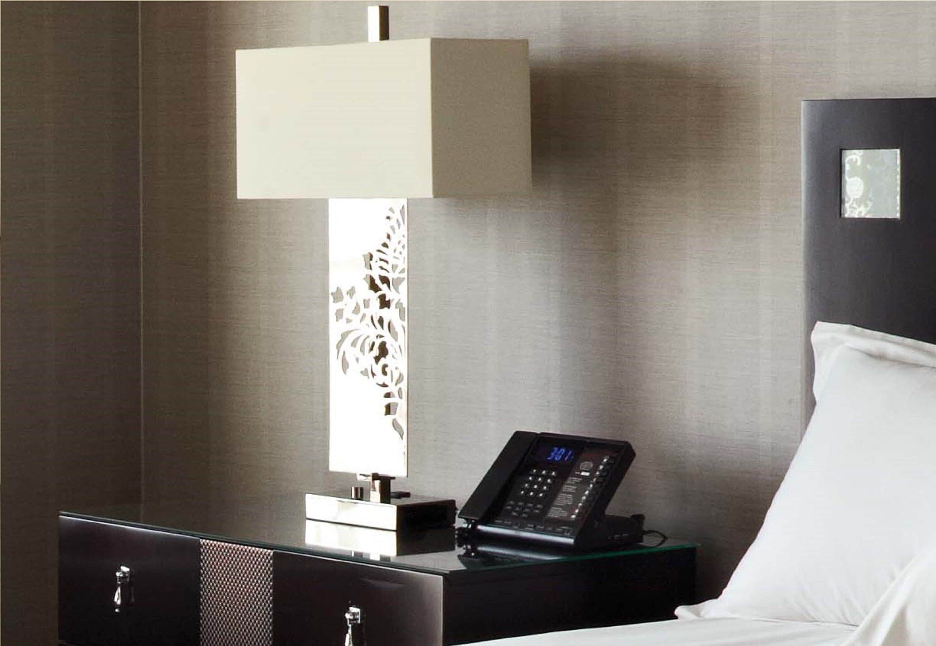 A hotel room with a bed, a bedside table with a lamp, a digital clock, and a small nightstand.