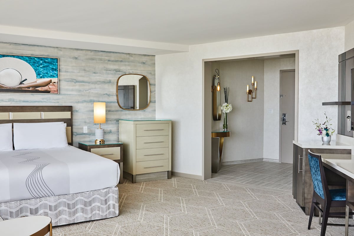 A hotel room with a bed, bedside table, lamp, dresser, mirror, and a desk with a chair.