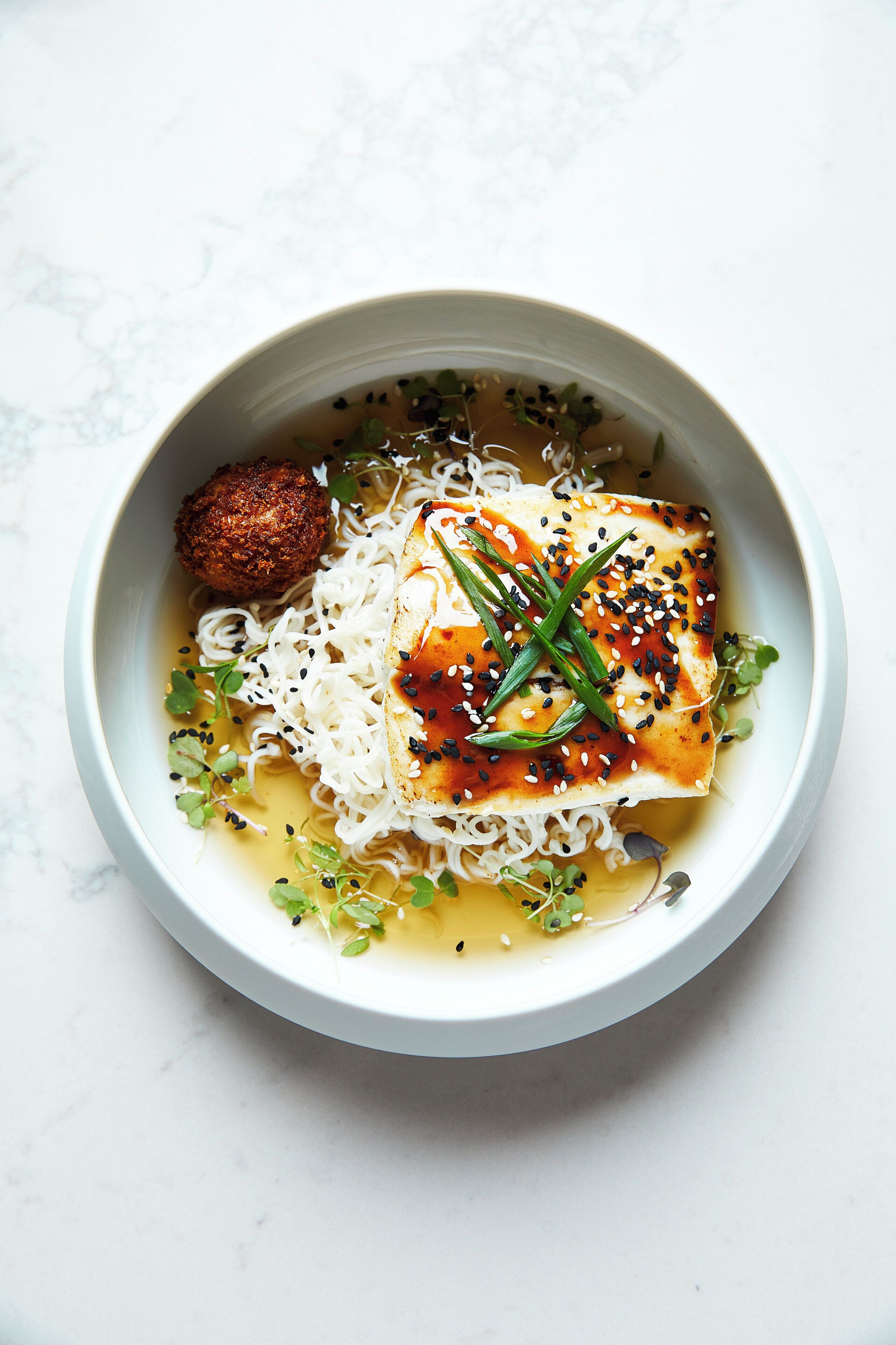 A bowl of ramen with a piece of fish, noodles, and garnishes.
