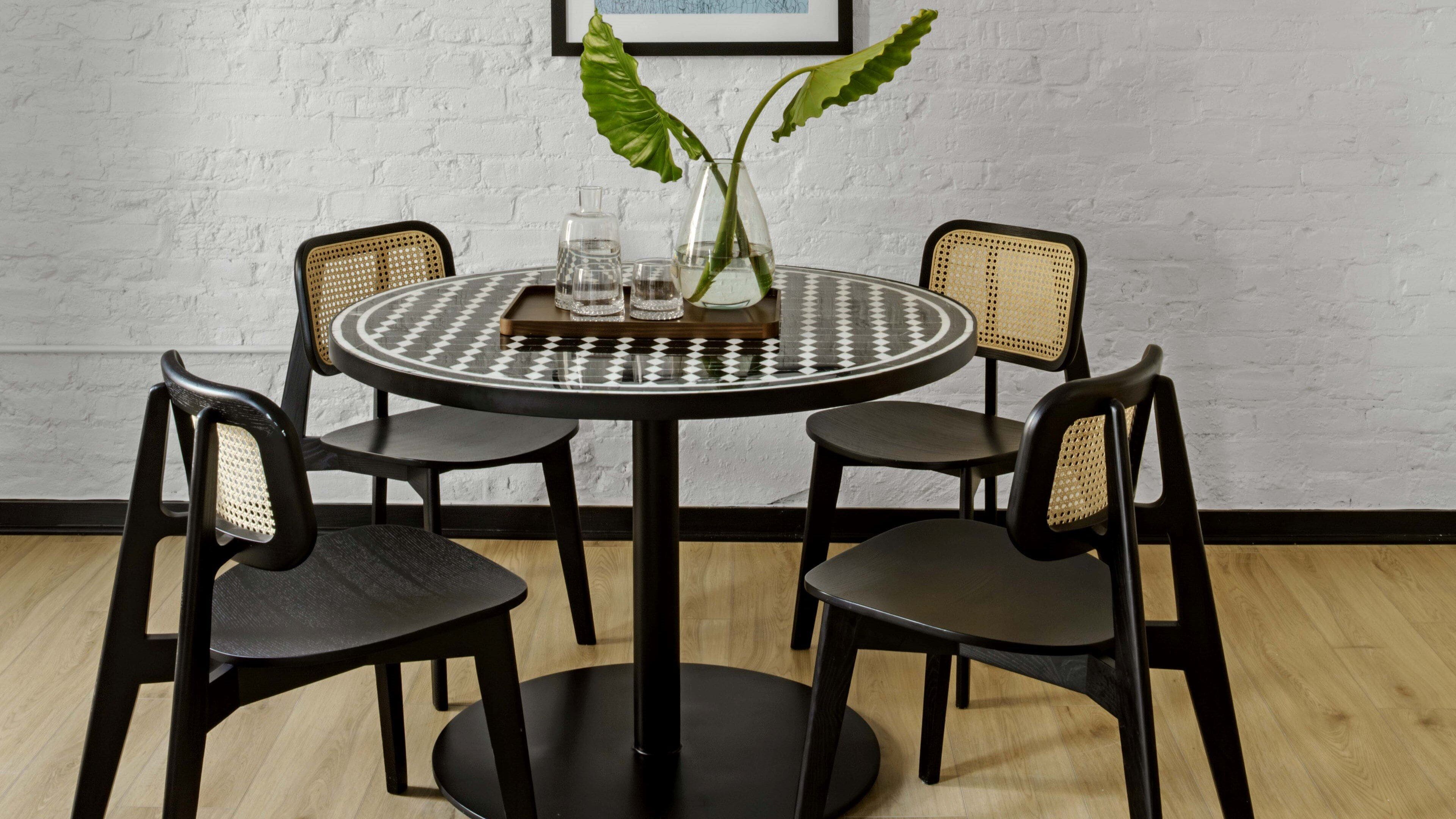 Une table ronde recouverte d’une nappe à carreaux noirs et blancs, entourée de quatre chaises noires dont les dossiers sont en osier. Un vase en verre contenant deux feuilles vertes est posé sur la table.