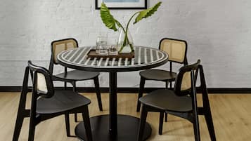 Una mesa redonda con mantel a cuadros blanco y negro, rodeada por cuatro sillas negras con respaldo trenzado. Un florero de vidrio con dos hojas verdes está sobre la mesa.