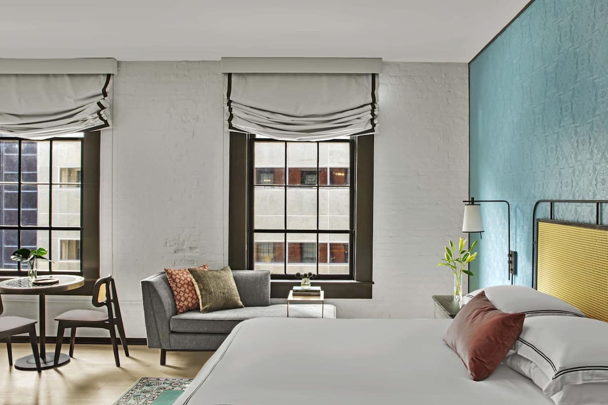 Ein modernes Hotelzimmer mit einem großen Bett, einer Couch, einem kleinen Tisch mit Stuhl und zwei Fenstern mit Vorhängen.