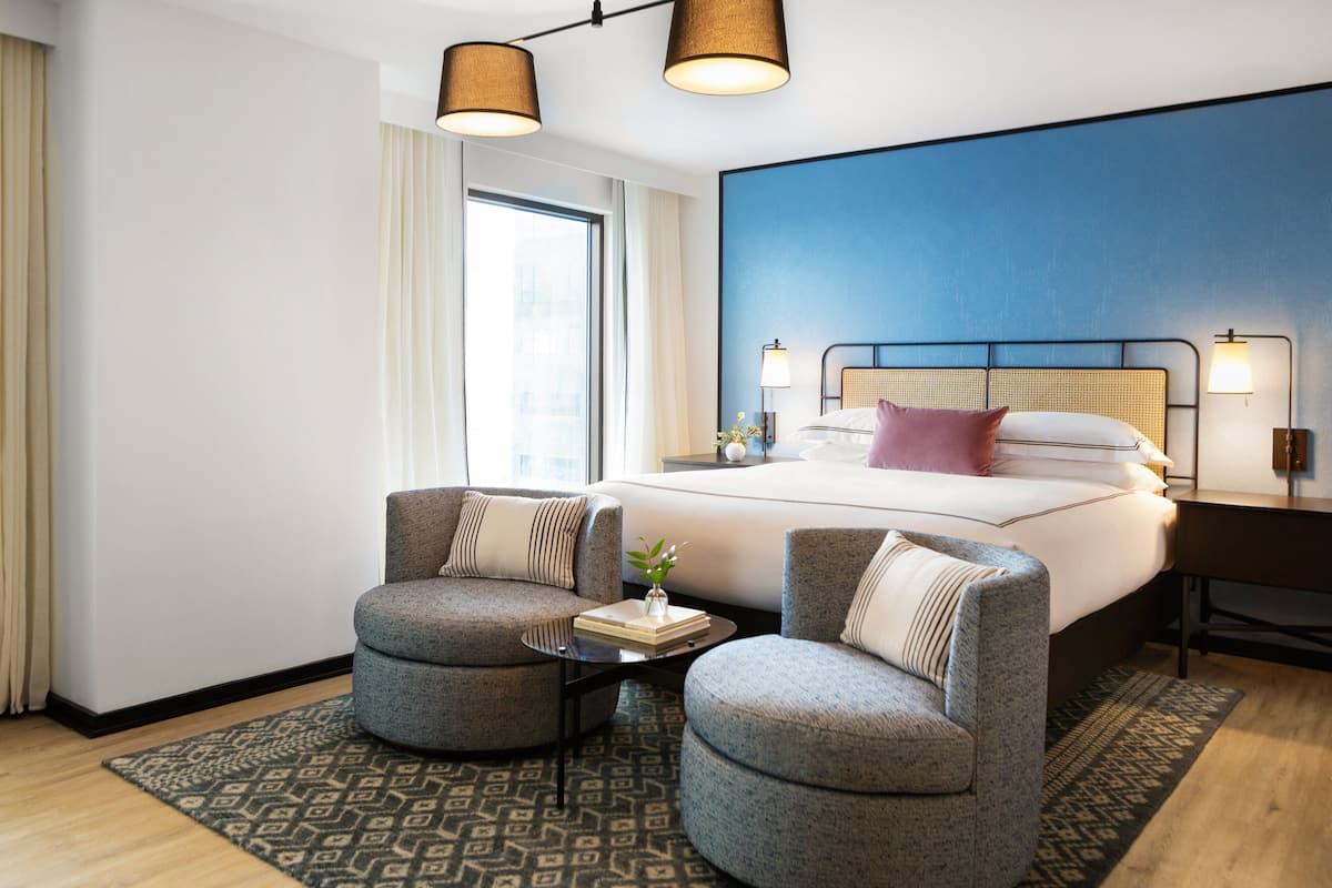Ein modernes Hotelzimmer mit einem großen Bett, zwei Sesseln, einem kleinen Tisch mit einem Buch und einer Vase sowie einem großen Fenster mit Vorhängen.