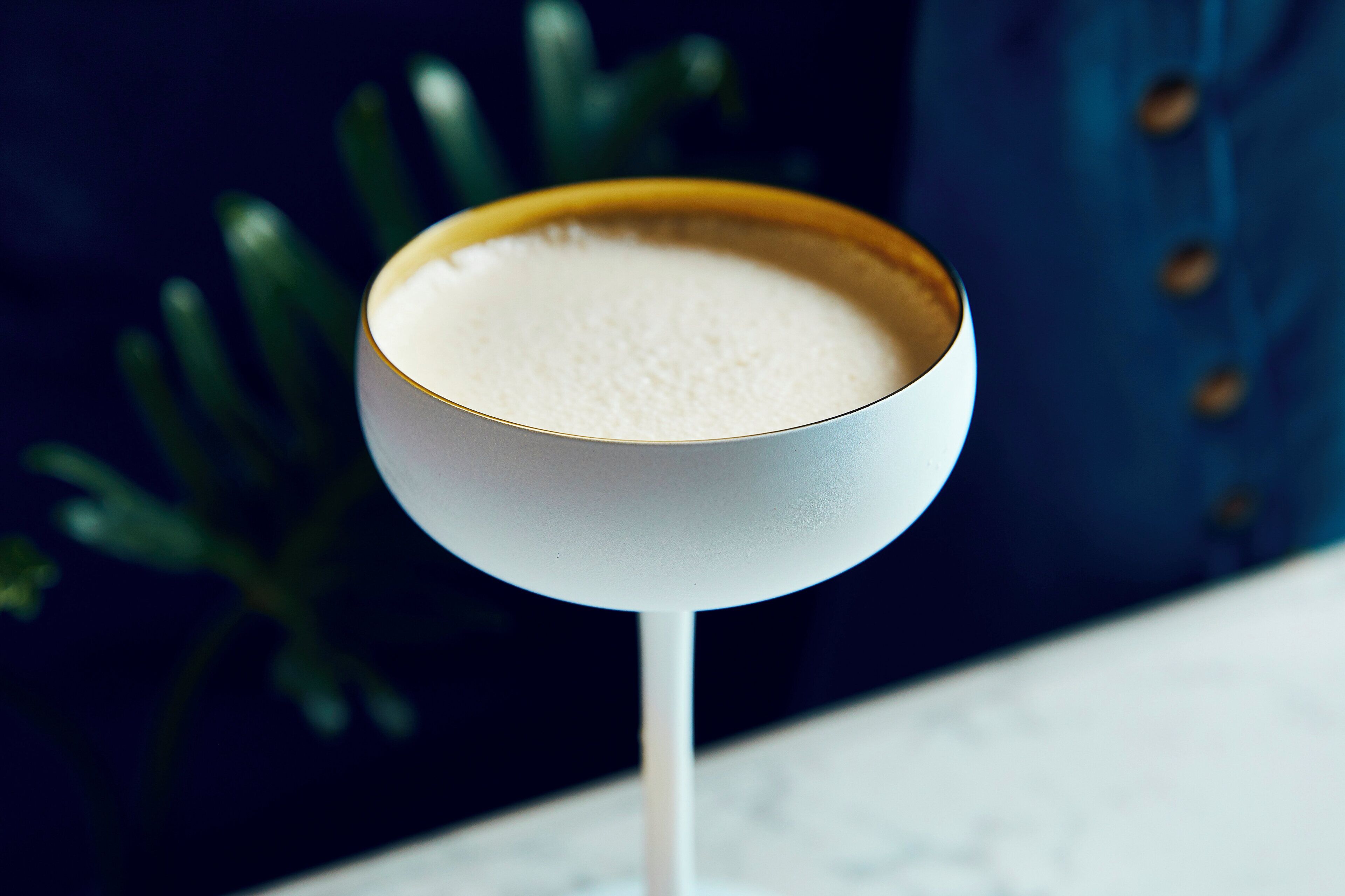 Un vaso de una bebida cremosa con espuma, sobre una mesa con fondo oscuro y una planta verde.