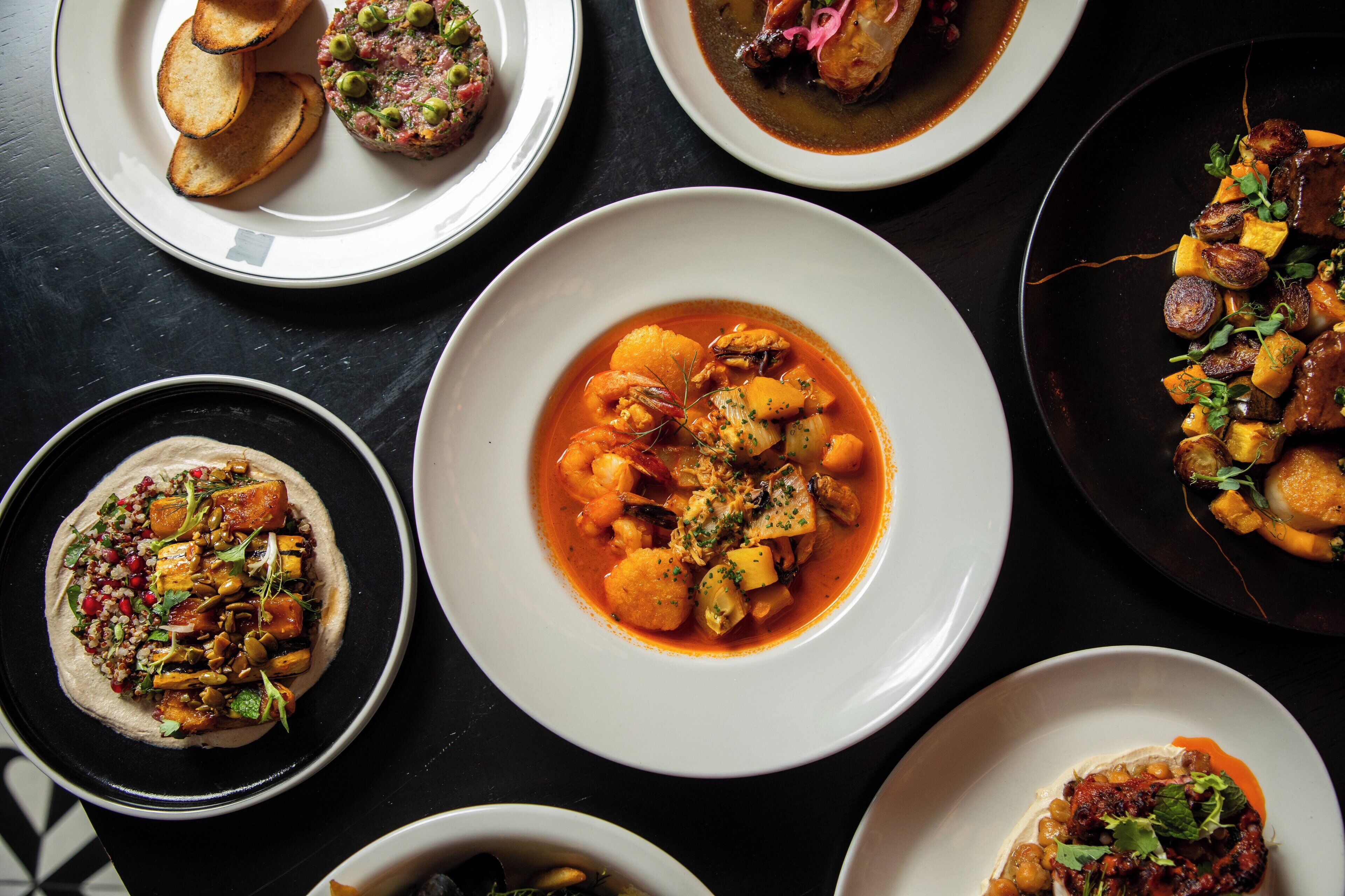 A variety of dishes on plates, including seafood, vegetables, and possibly some meat, arranged on a dark surface.