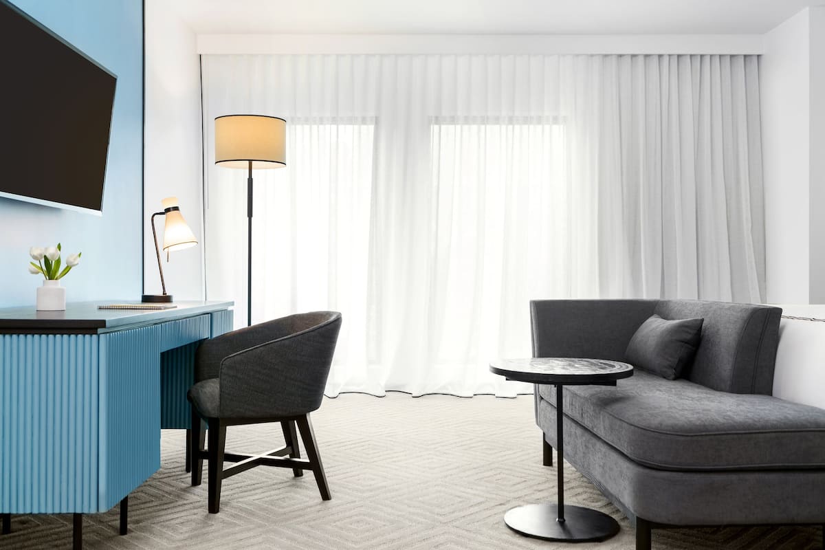 A modern hotel room with a blue desk, a grey sofa, a small round table, and a floor lamp.