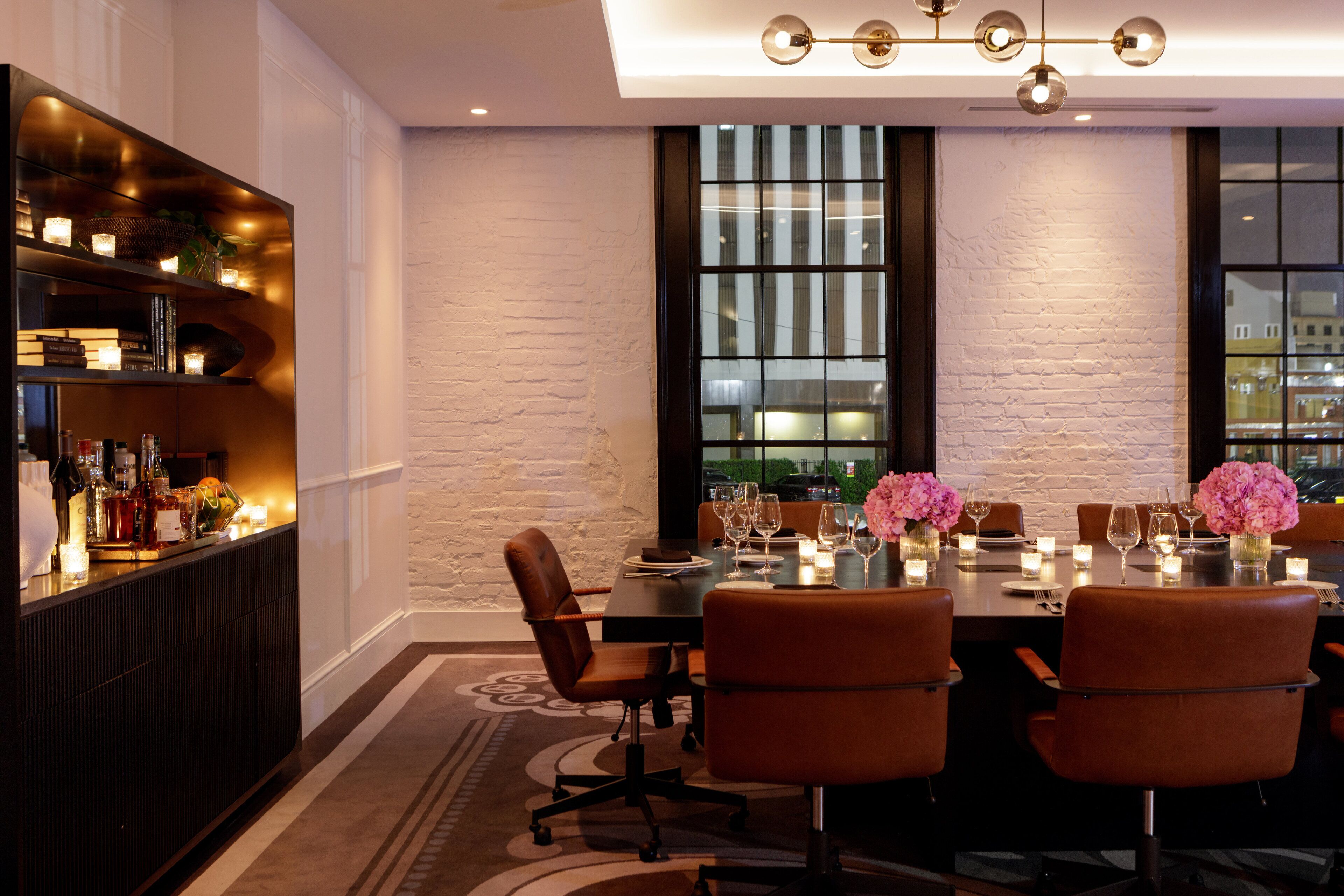 Un espace repas avec une longue table dressée pour six personnes, entourée de fauteuils en cuir, et un coin bar à gauche, ainsi que de grandes fenêtres offrant une vue sur le paysage urbain.