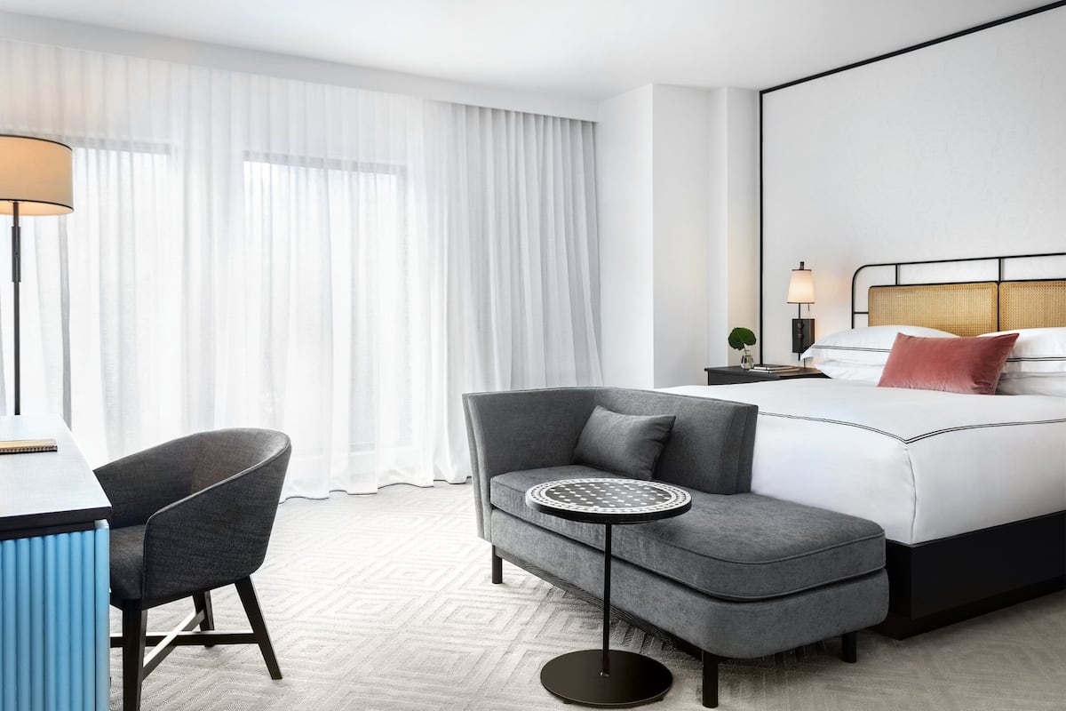 A hotel room with a bed, a desk, a chair, a small table, and a lamp.