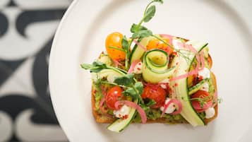 A slice of toast topped with avocado, cherry tomatoes, cucumber, and herbs on a white plate.