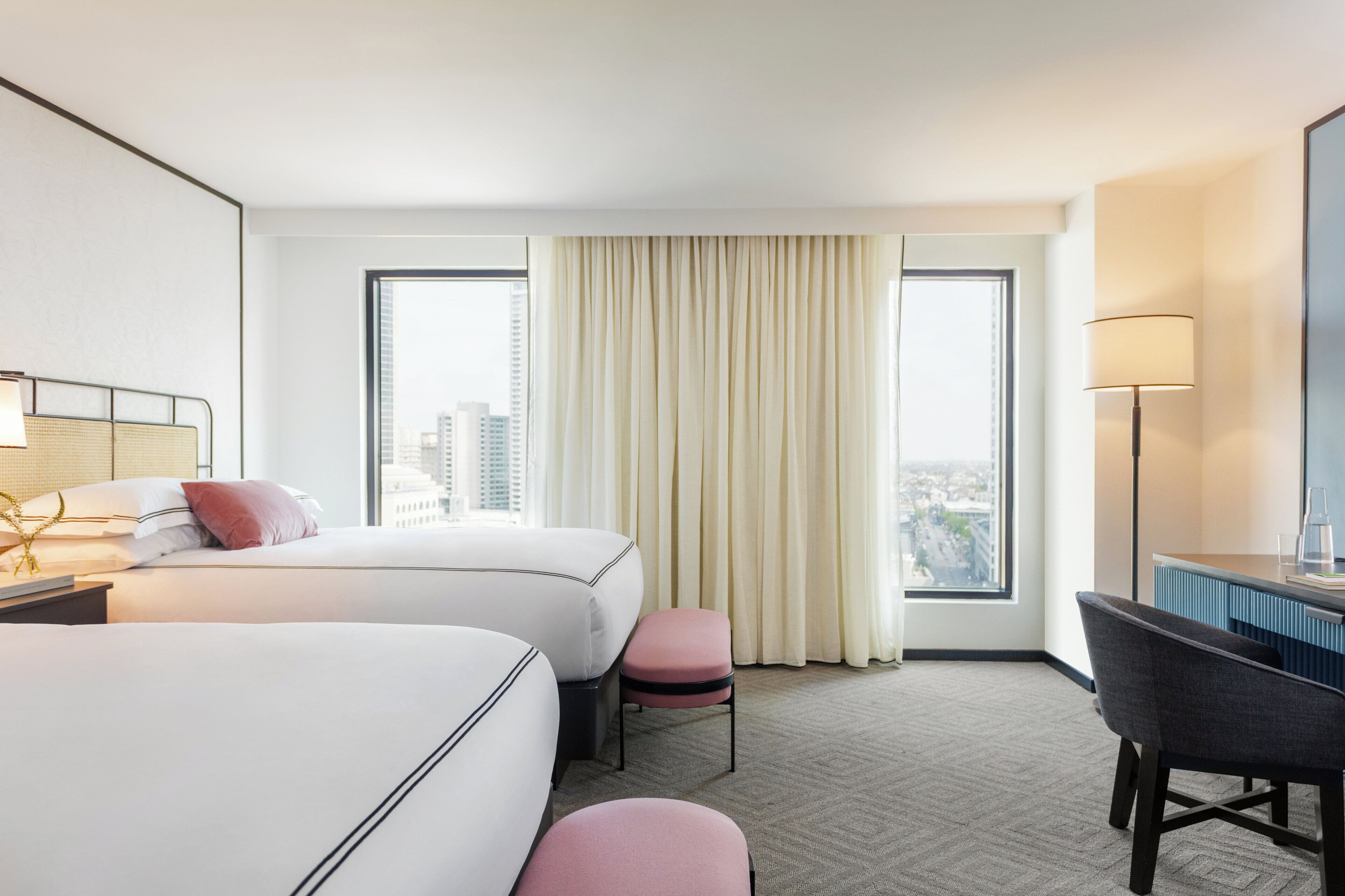 A hotel room with two beds, a desk, a chair, and a large window with curtains.