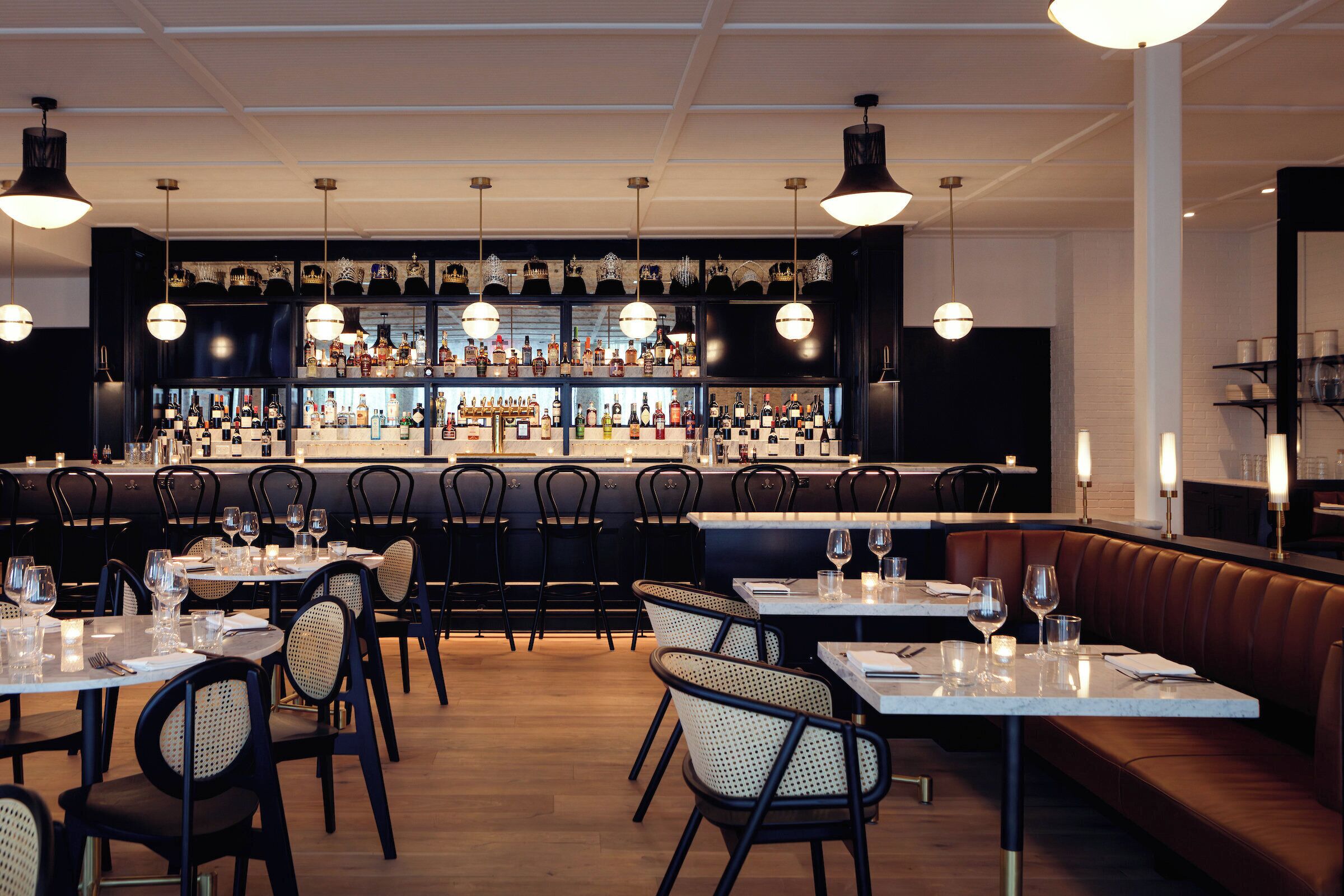 A restaurant with multiple tables and chairs, a bar area with shelves of bottles, and pendant lighting.