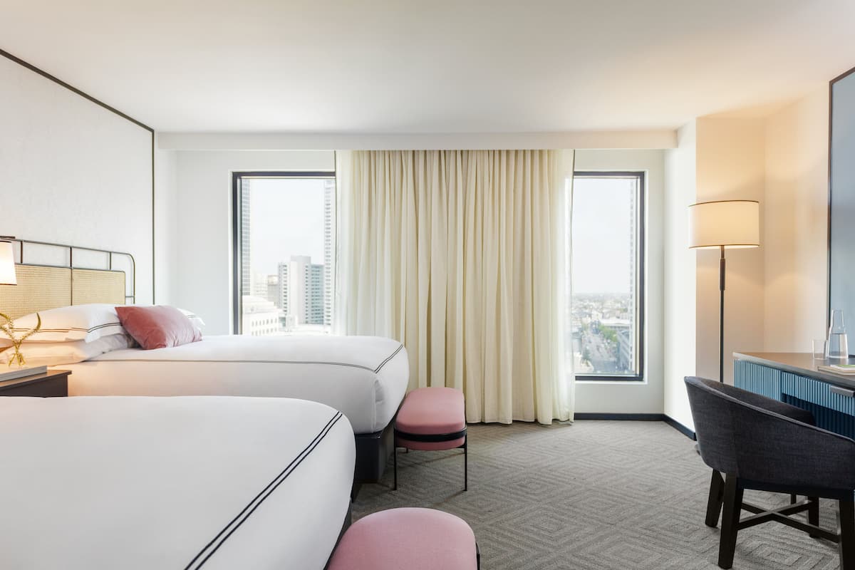 Ein Hotelzimmer mit zwei Betten, einem Schreibtisch, einem Stuhl, einer Lampe und Blick auf die Stadt durch große Fenster.