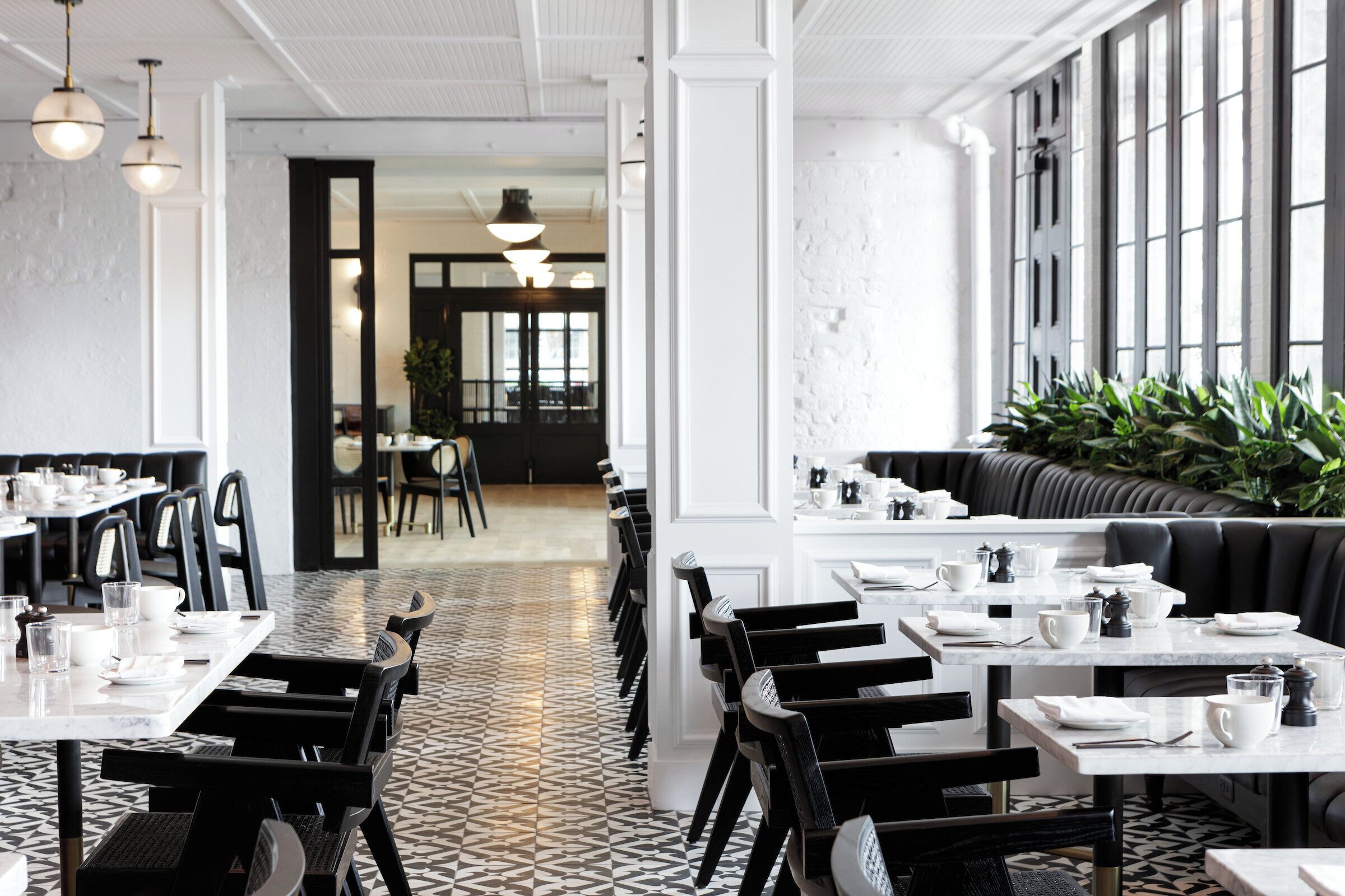 A modern restaurant with black and white tiled flooring, black chairs, and white tables. There are large windows on one side and a plant in the corner.
