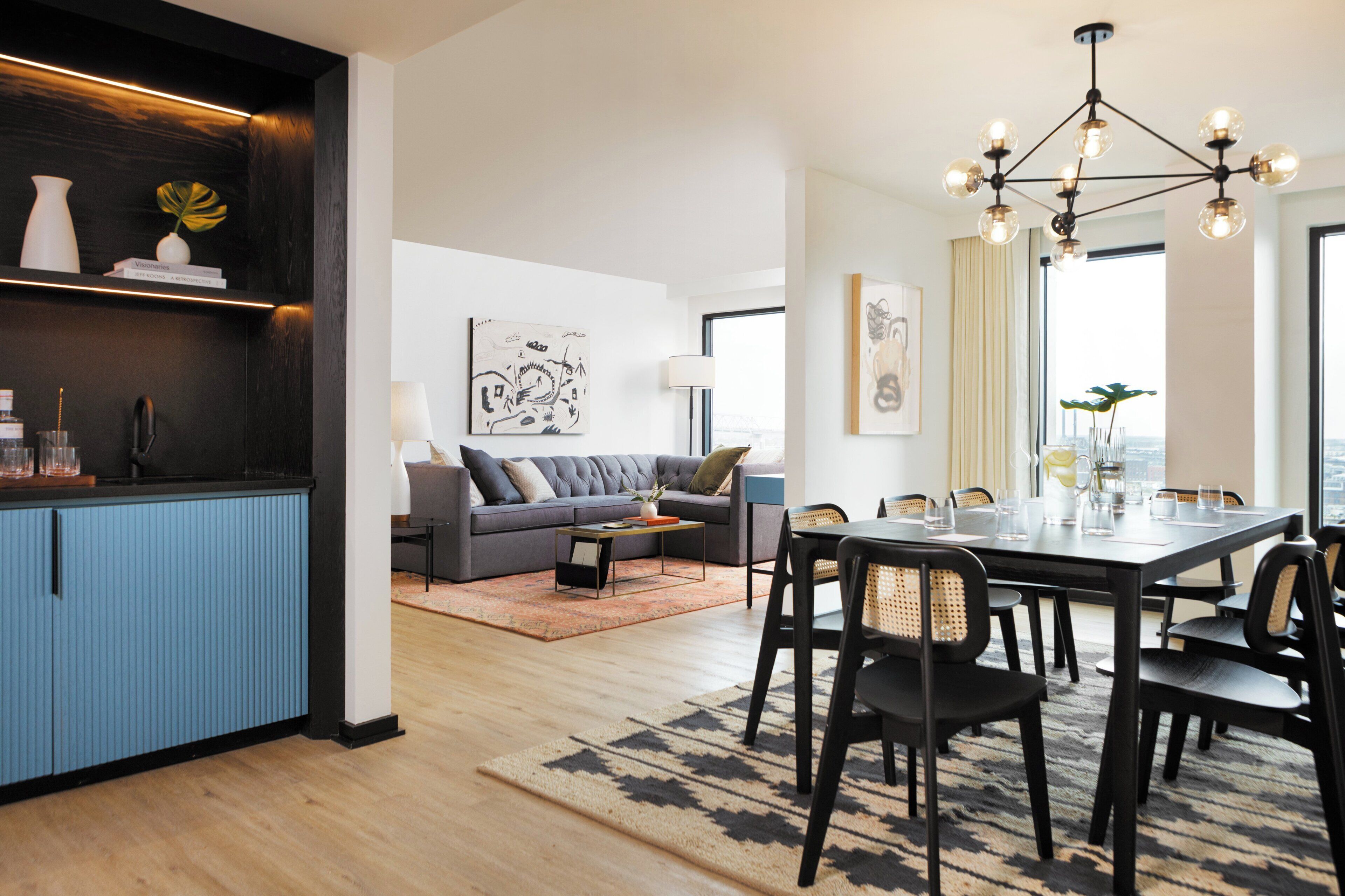 Un salon moderne avec un canapé gris, une table à manger avec des chaises, un coin cuisine avec des meubles bleus et des œuvres d’art accrochées aux murs.