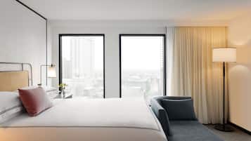 A hotel room with a large bed, a desk, a chair, a floor lamp, and a view of the city through two large windows.
