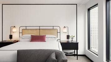 A modern hotel room with a large bed, two bedside lamps, a small desk, and a large window with a city view.