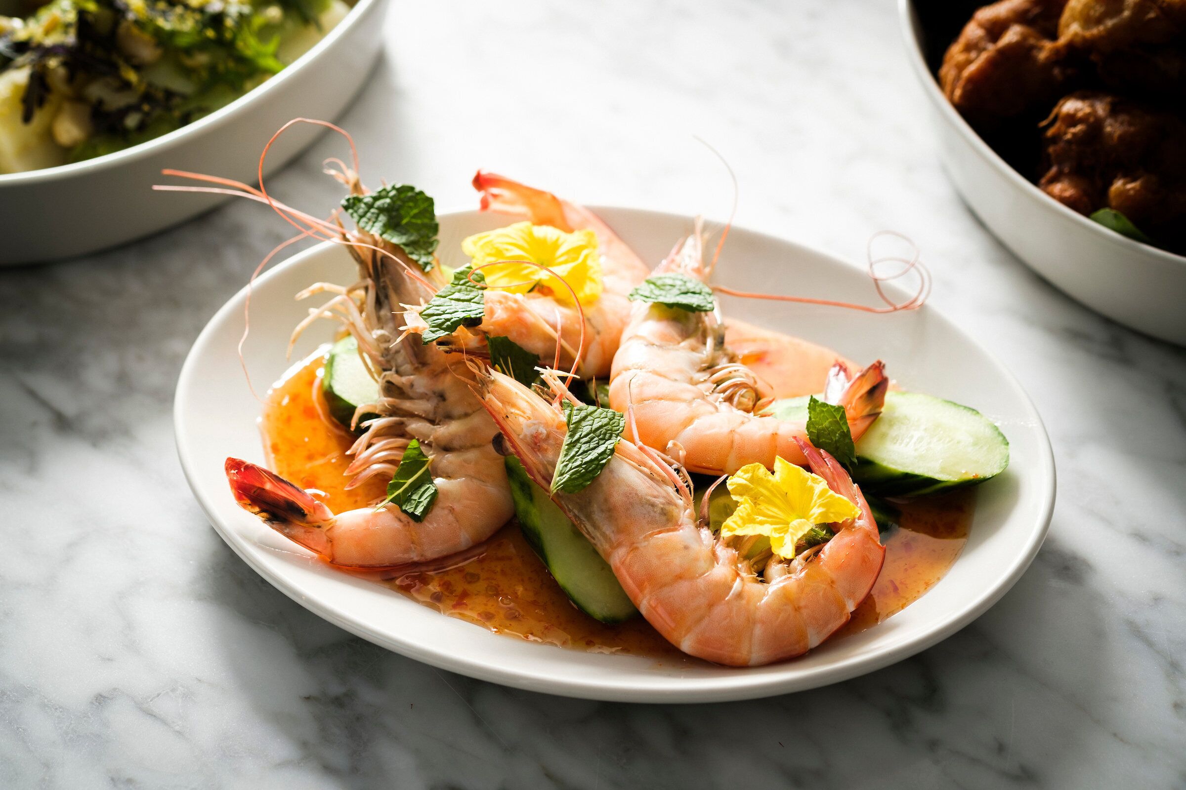 A plate of cooked shrimp garnished with mint leaves and a yellow flower, served on a white plate.
