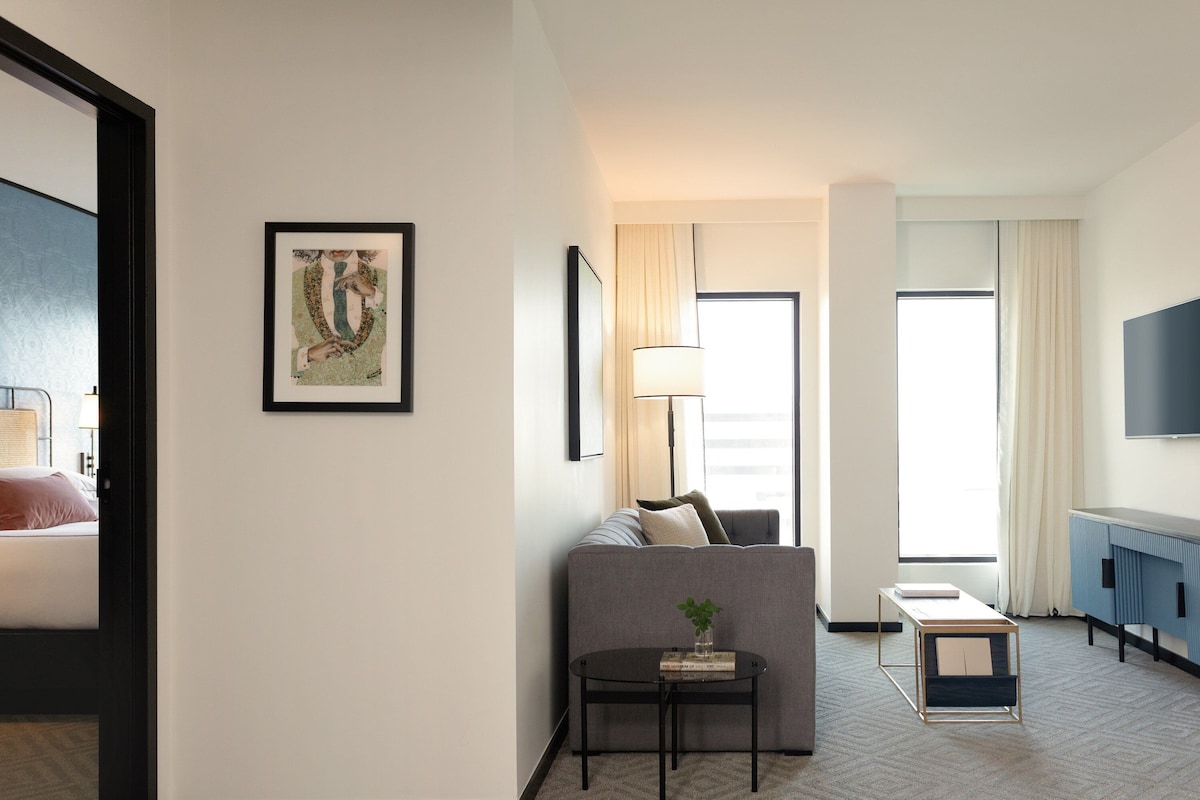Una habitación de hotel moderna con sofá, una mesita con una planta, una mesita de noche, una lámpara, un televisor y una ventana con cortinas.