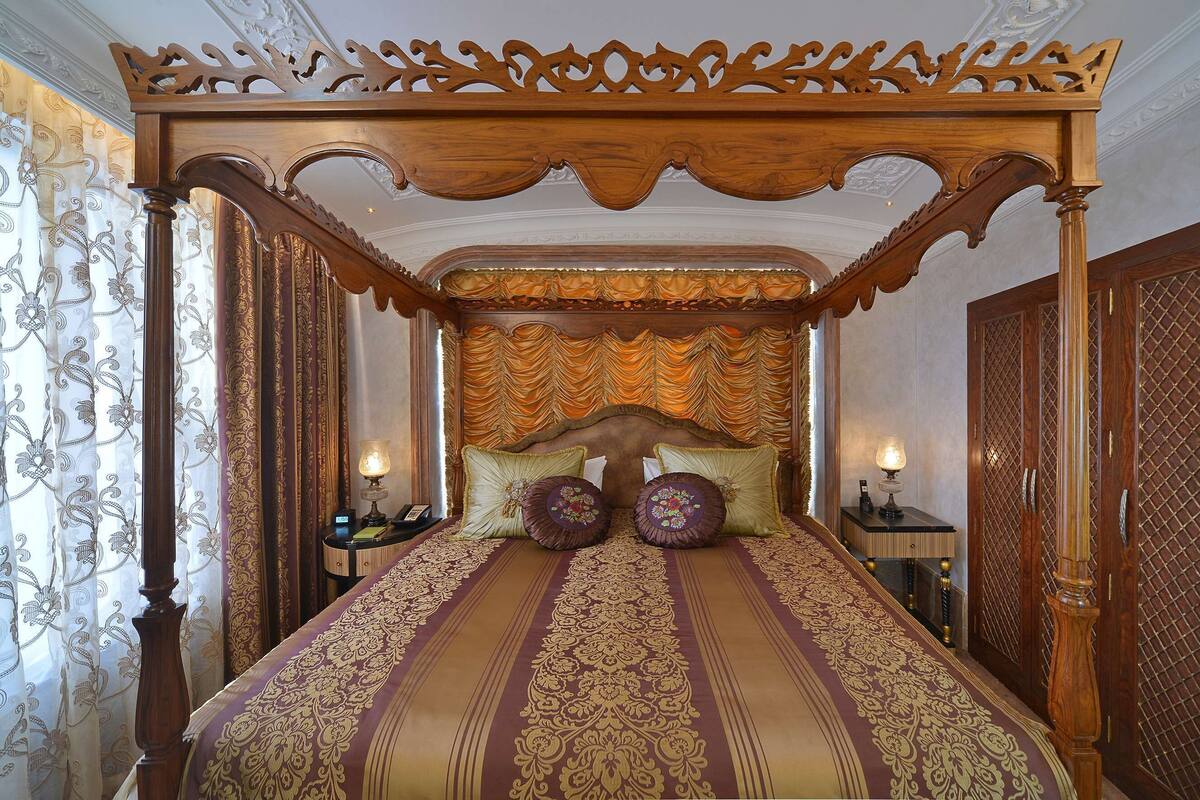 A four-poster bed with ornate headboard and curtains, a nightstand with a lamp, and a wooden wardrobe.