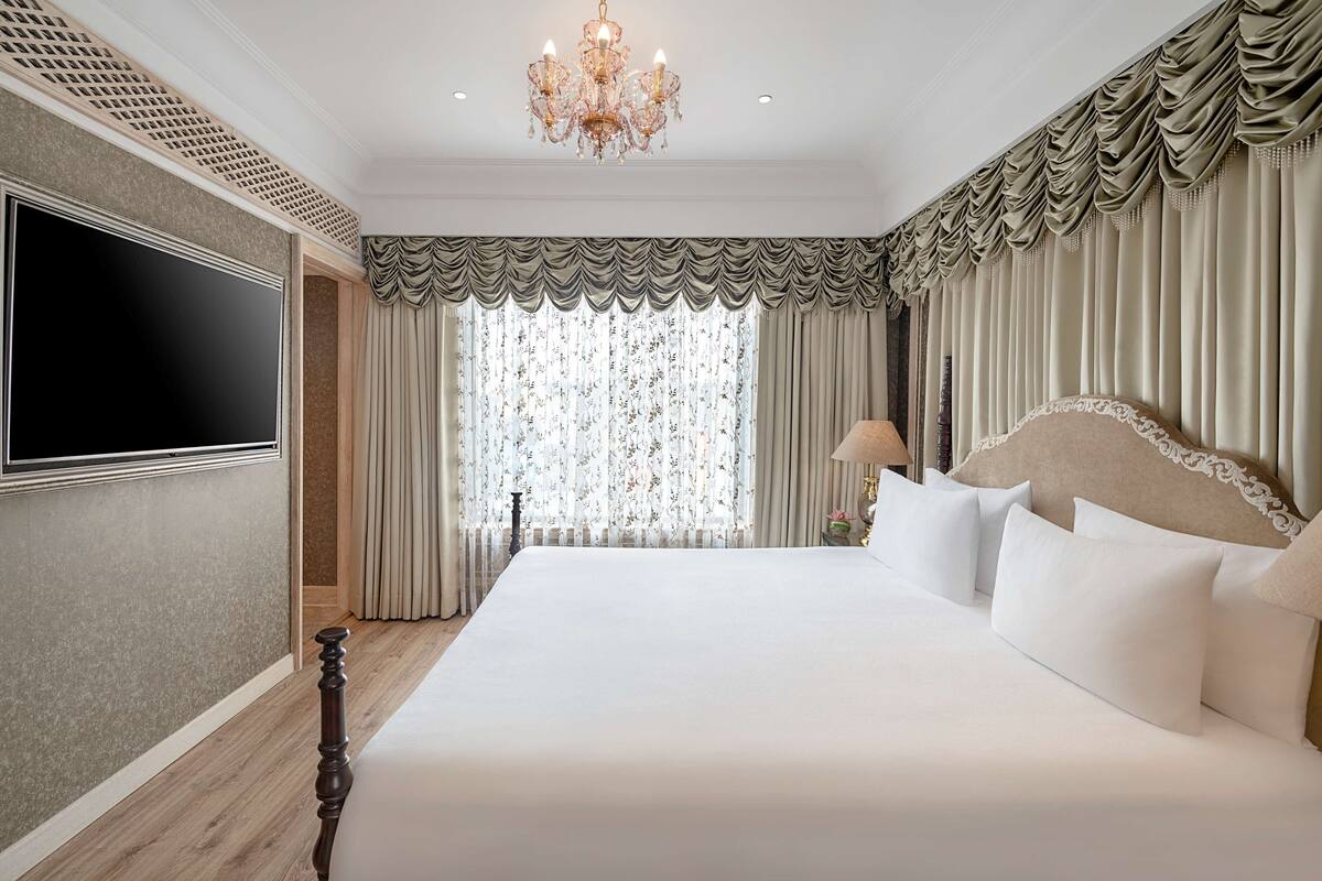 A spacious bedroom with a large bed, a flat-screen TV mounted on the wall, and a chandelier.