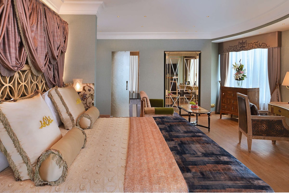 A spacious hotel room with a large bed, a sitting area with a sofa and armchairs, a wooden desk, and a television.