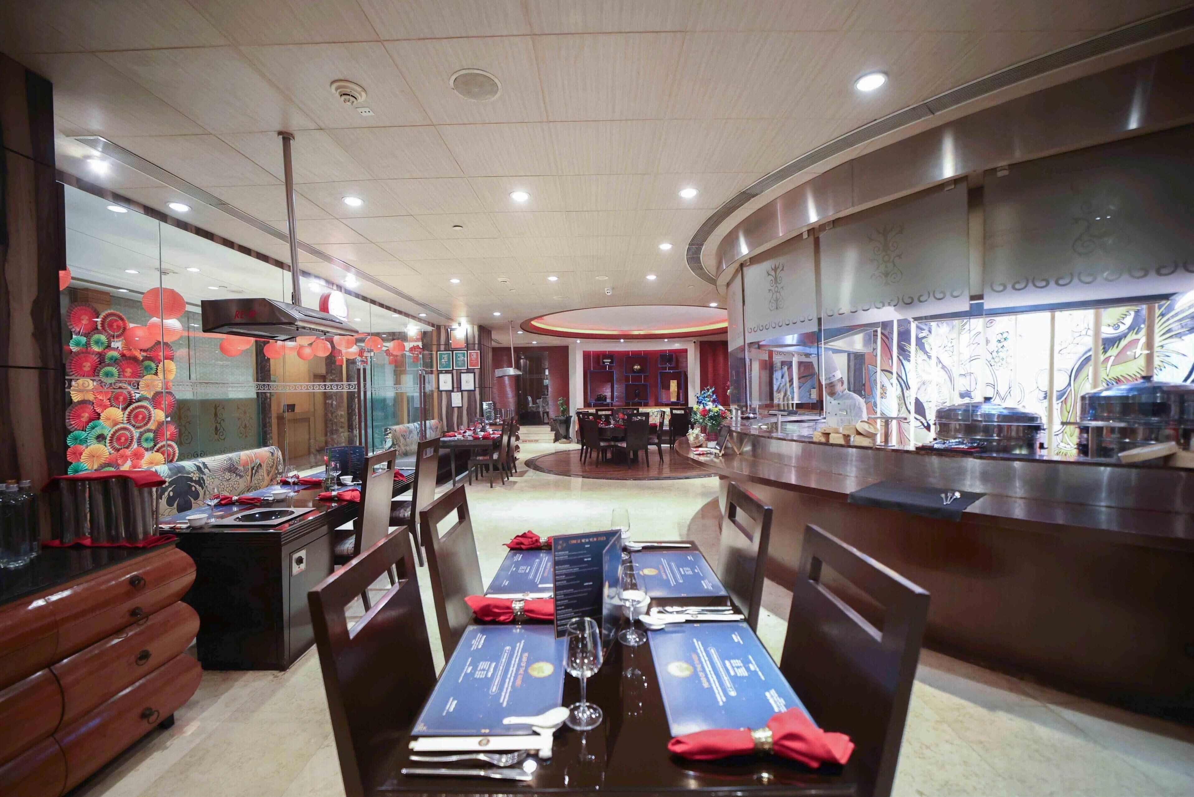 A restaurant with a central dining area featuring a long table set for two, surrounded by chairs. There is a counter on the right with a display of food items and a person working behind it. The ceiling has recessed lighting, and the walls have decorative panels.
