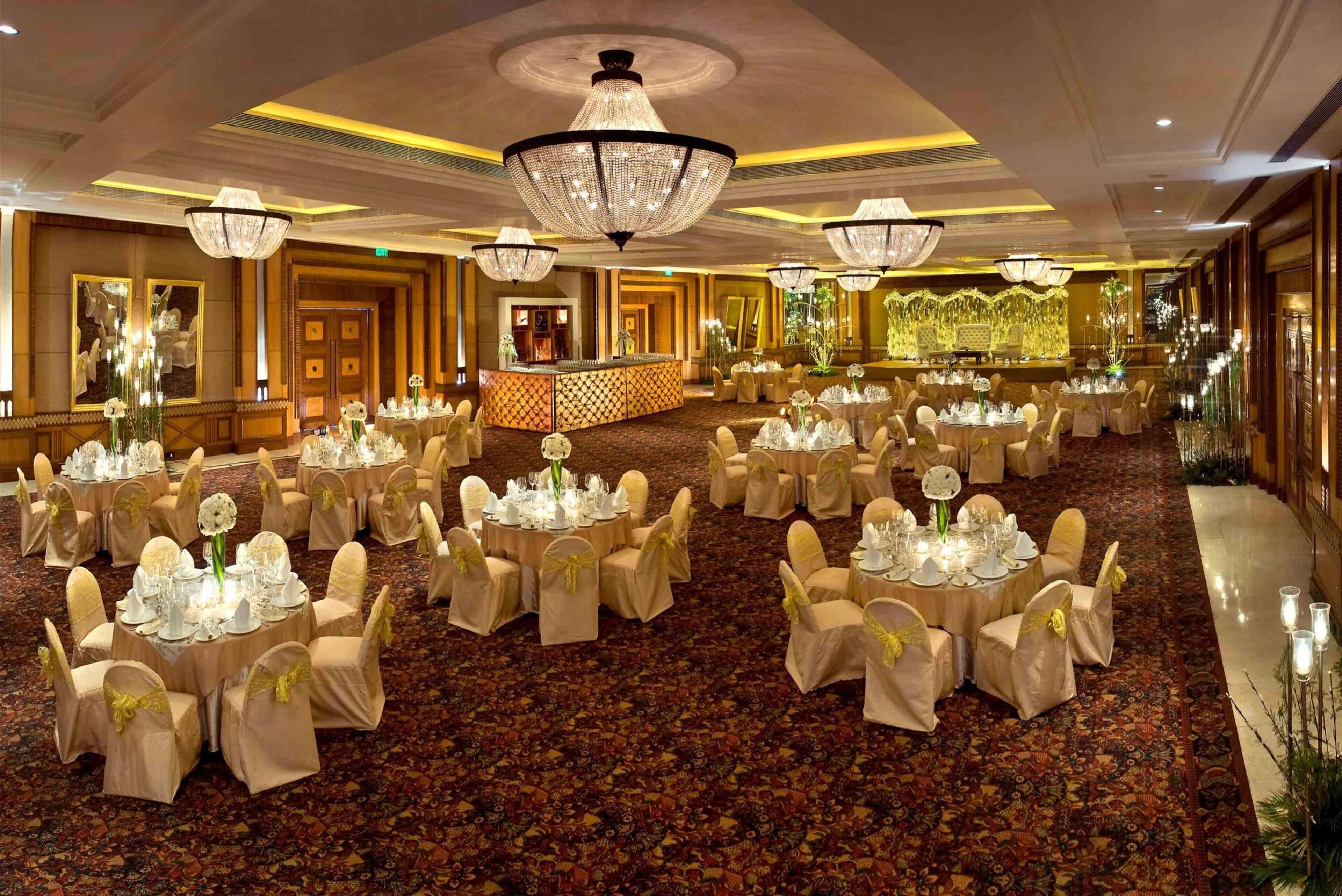 A banquet hall with round tables set for events, featuring chandeliers and a carpeted floor.