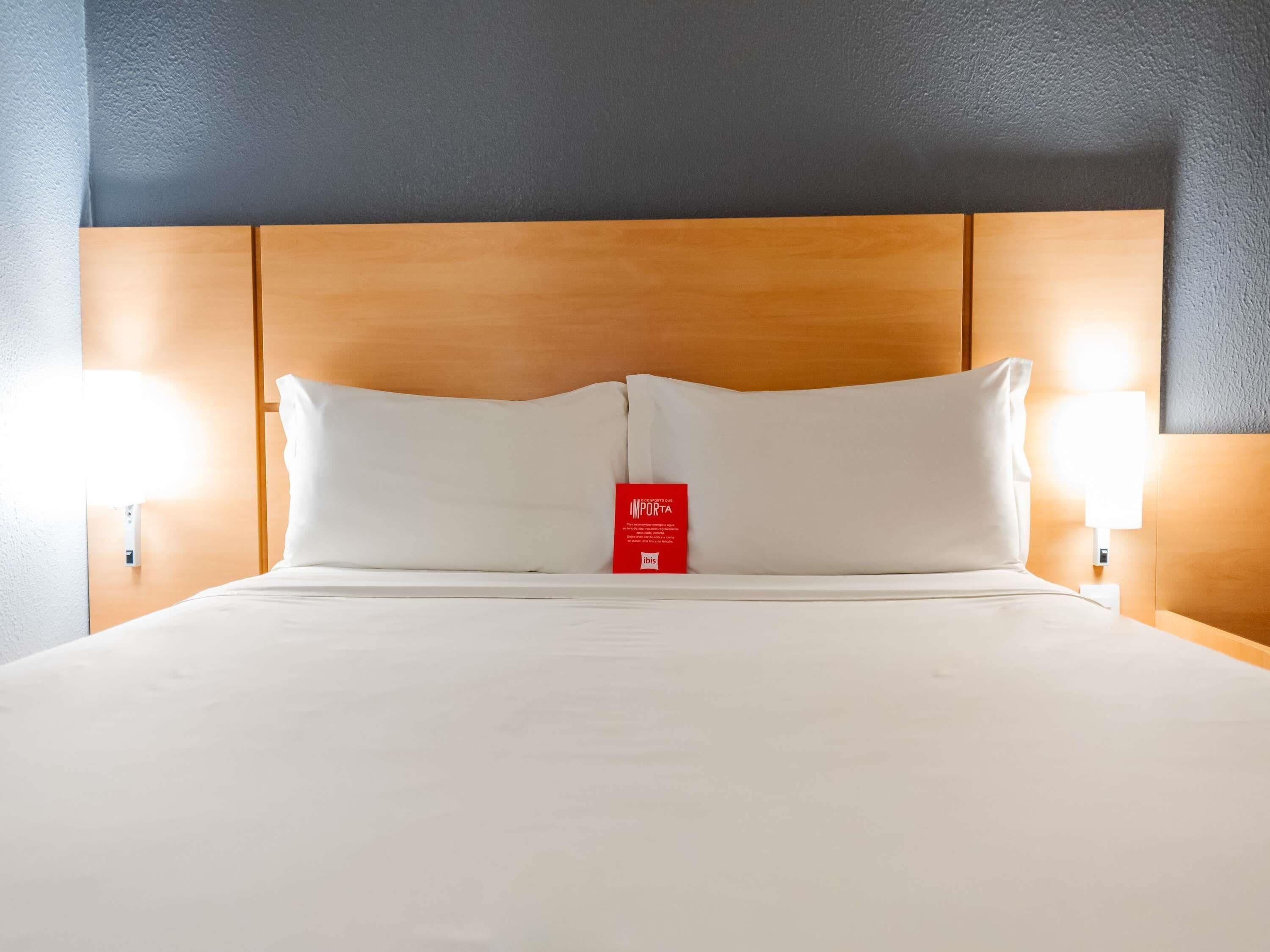 Quarto de hotel com cabeceira de madeira, duas almofadas brancas, cartão de minibar vermelho e luminárias fixadas na parede.