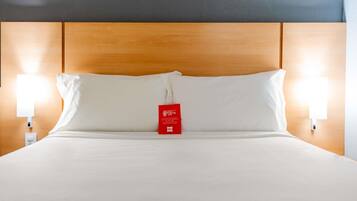 Une chambre d’hôtel avec une tête de lit en bois, du linge de lit blanc, deux lampes de chevet et une étiquette rouge sur le lit.