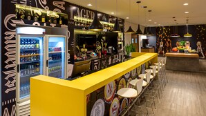 A bar area with a yellow counter, multiple stools, and a refrigerator stocked with beverages. - ibis Joinville (Joinville)