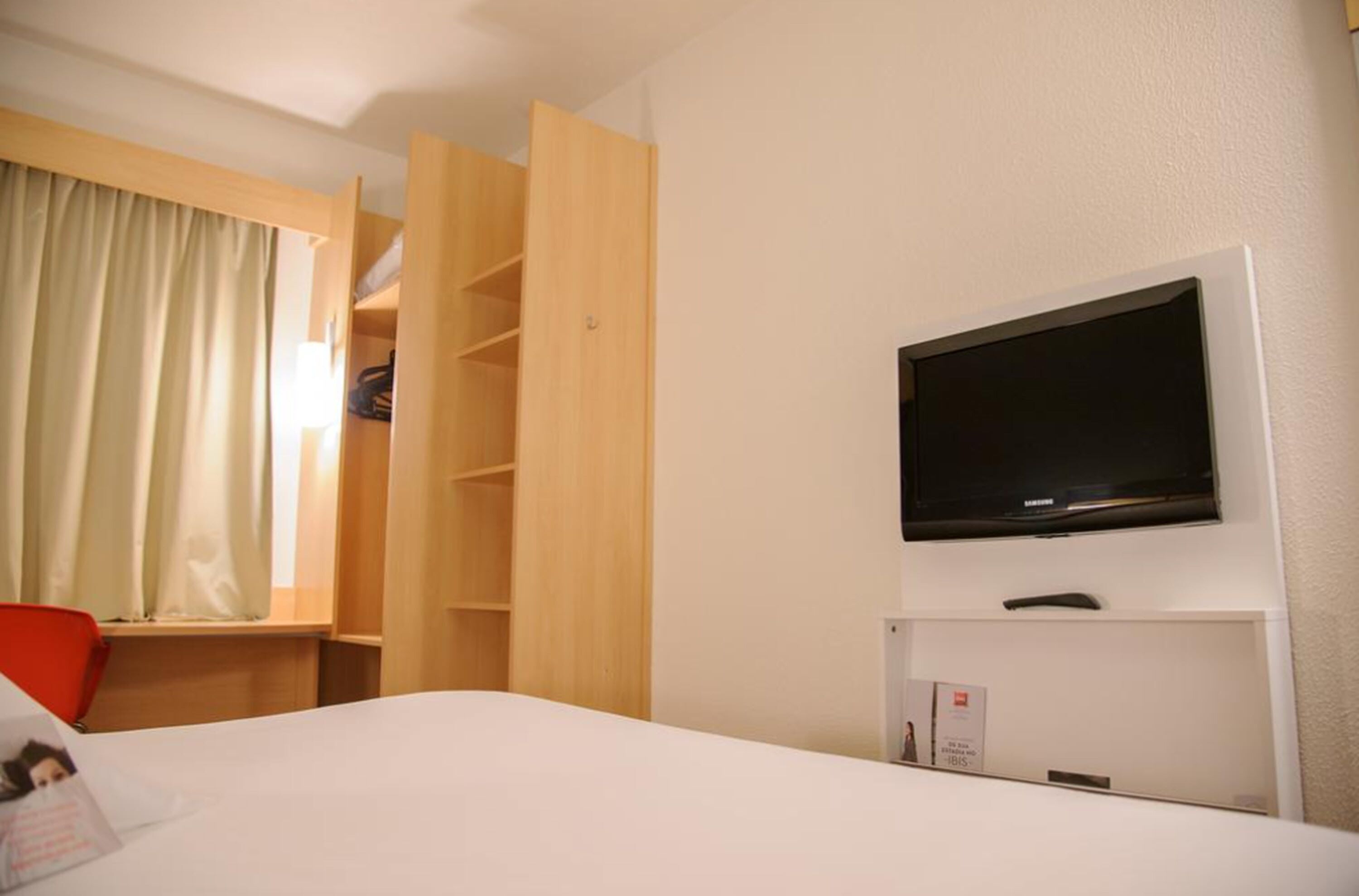 A hotel room with a bed, a TV mounted on the wall, a wooden wardrobe, and a window with curtains.