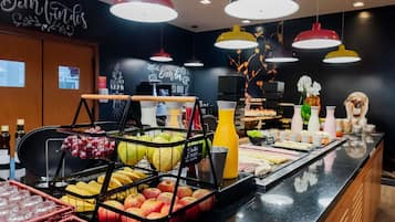 Una barra de desayuno con diversas frutas, un dispensador de jugo y un menú en una pizarra.