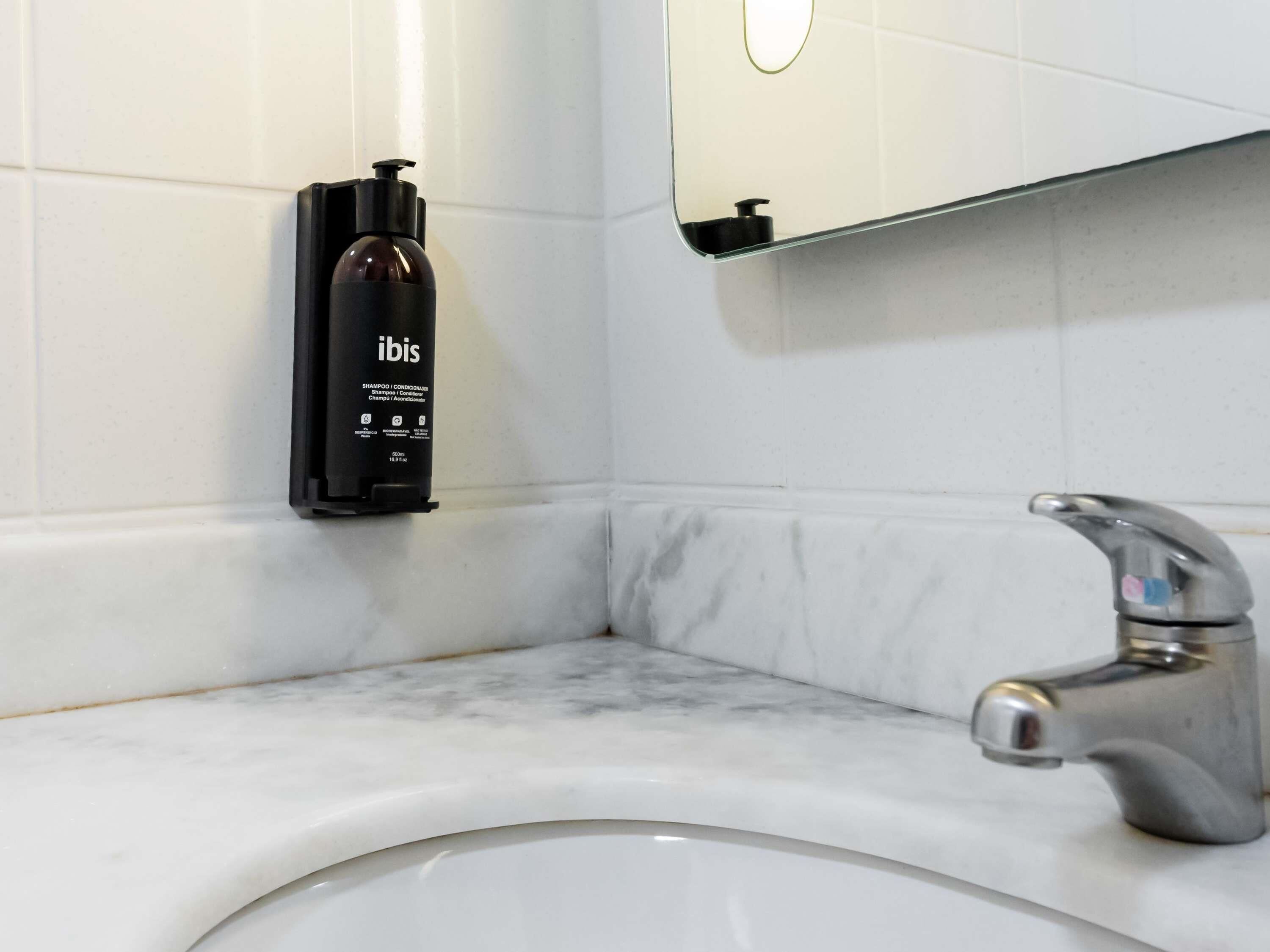 Un baño con un lavamanos de mármol, un grifo moderno y una botella negra de champú con la etiqueta 'ibis' en la pared.