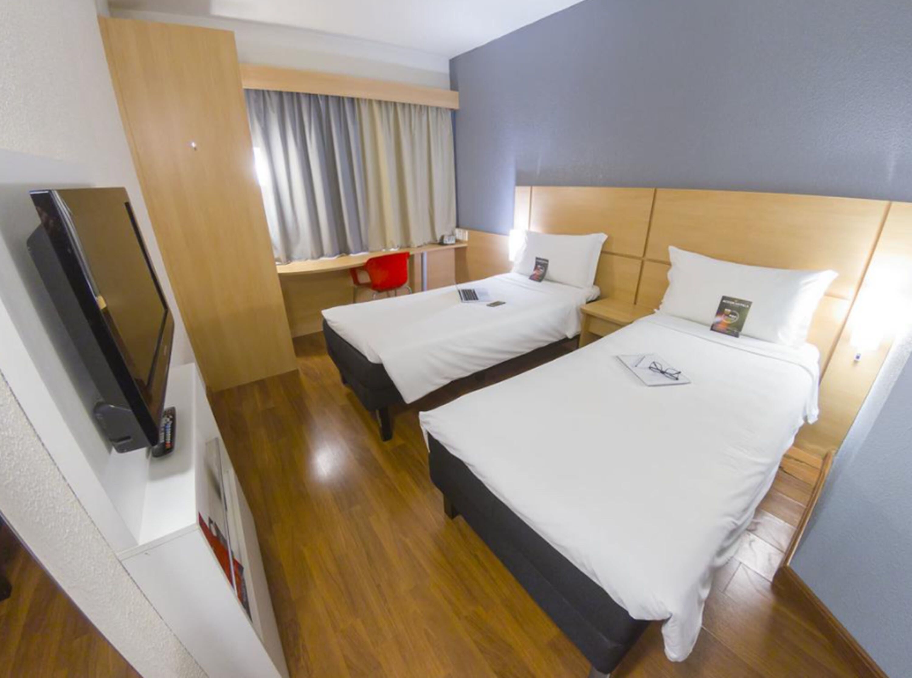 A hotel room with two beds, a desk, a TV, and a window with curtains.