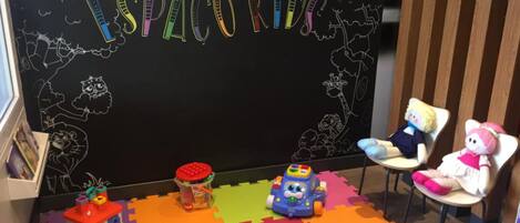 A children's play area with a blackboard titled "ESPAZO KID," featuring drawings of animals and clouds. There are colorful foam mats, a toy car, and a toy truck on the floor.
