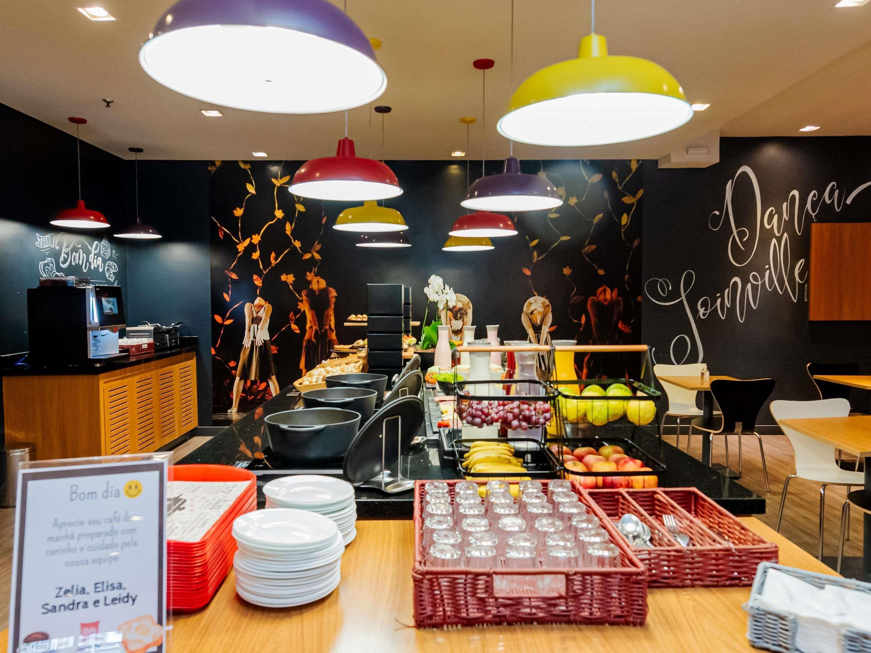 Un café con pared negra decorada con un mural de un árbol frutal, una barra con un refrigerador y una variedad de frutas y bebidas a la vista.