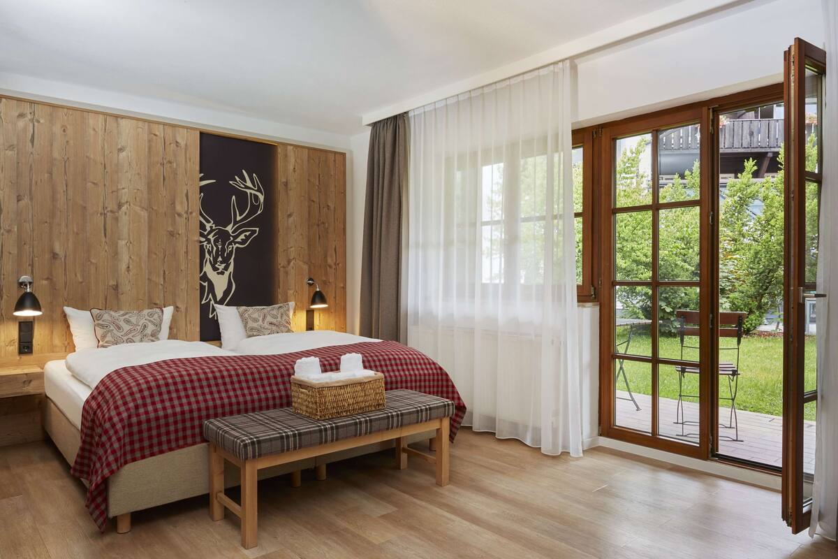 A bedroom with a large bed, a bench, a wicker basket, and a view of the outdoors through a glass door.