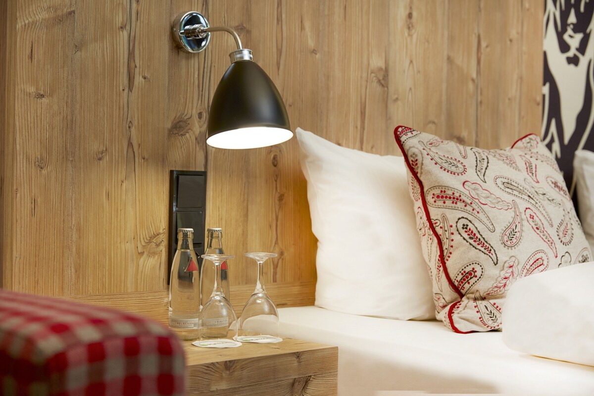 A wooden wall with a modern pendant light, a white sofa with patterned pillows, and a wooden nightstand with wine glasses.