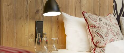 A wooden wall with a modern pendant light, a white sofa with patterned pillows, and a wooden nightstand with wine glasses.