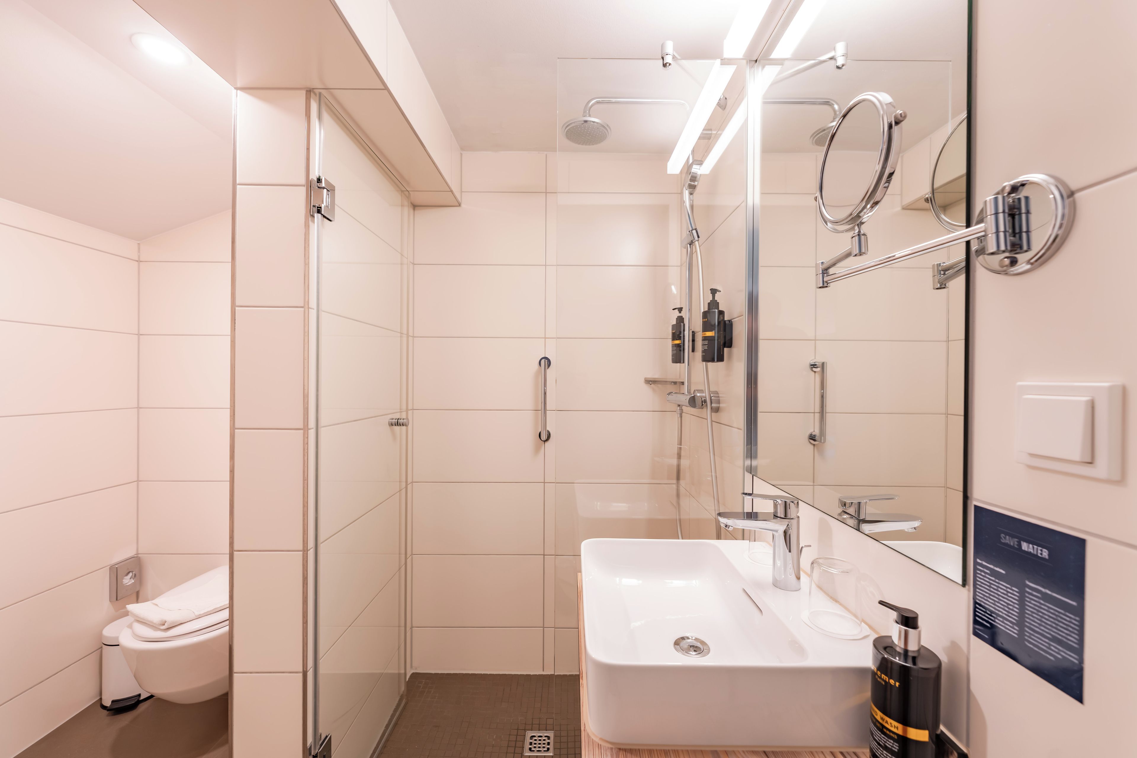 A modern bathroom with a glass shower enclosure, a white sink, and a mirror with a 'SAVE WATER' sign.