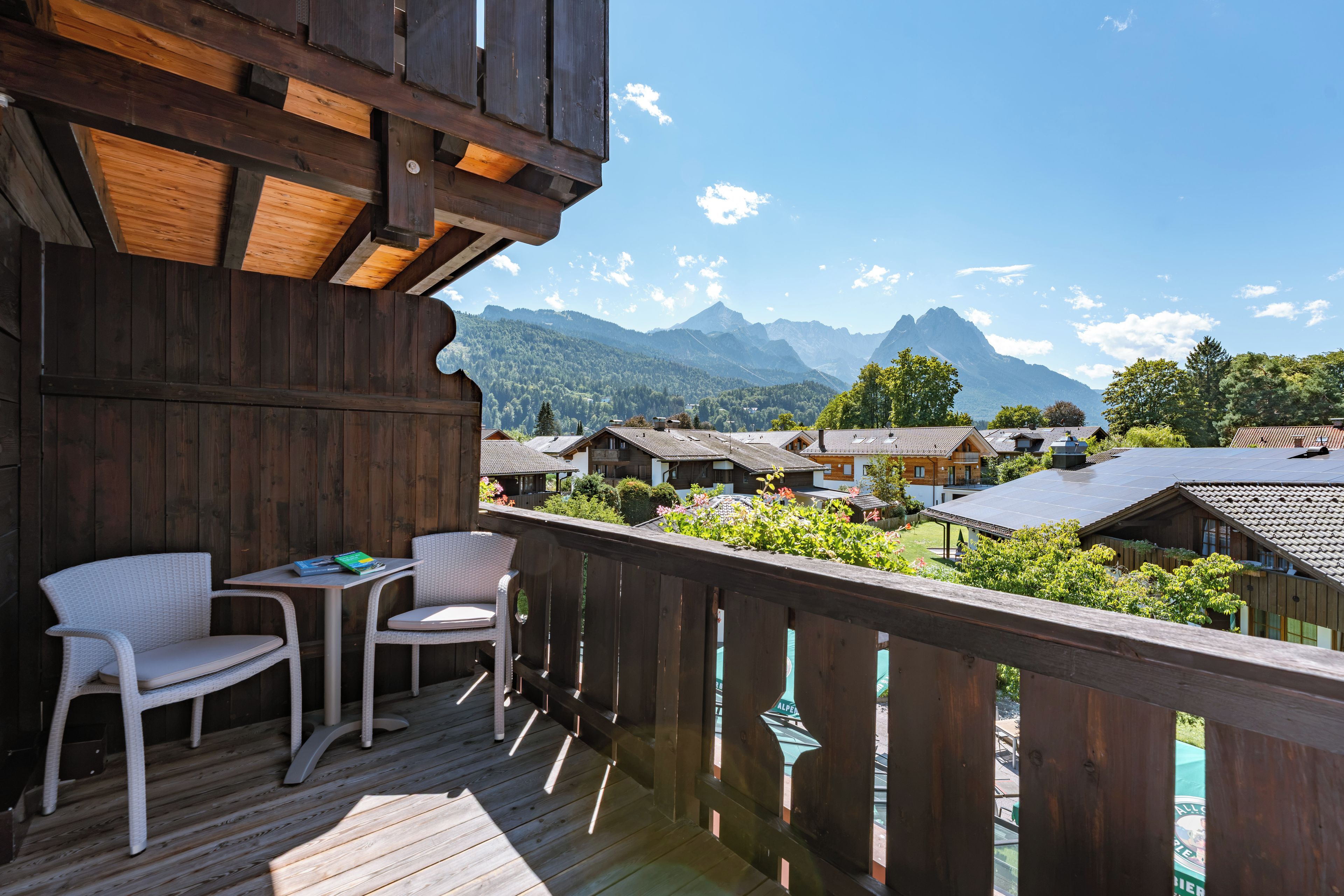 A wooden balcony with wicker furniture, overlooking a village with traditional houses and mountains in the background.