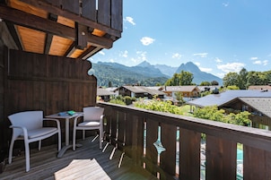 Un balcon en bois, agrémenté de meubles en osier, qui offre une vue sur un village aux maisons traditionnelles, avec des montagnes en toile de fond.