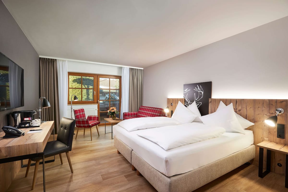 A hotel room with a bed, a desk with a telephone, a chair, a table with flowers, and a window with curtains.