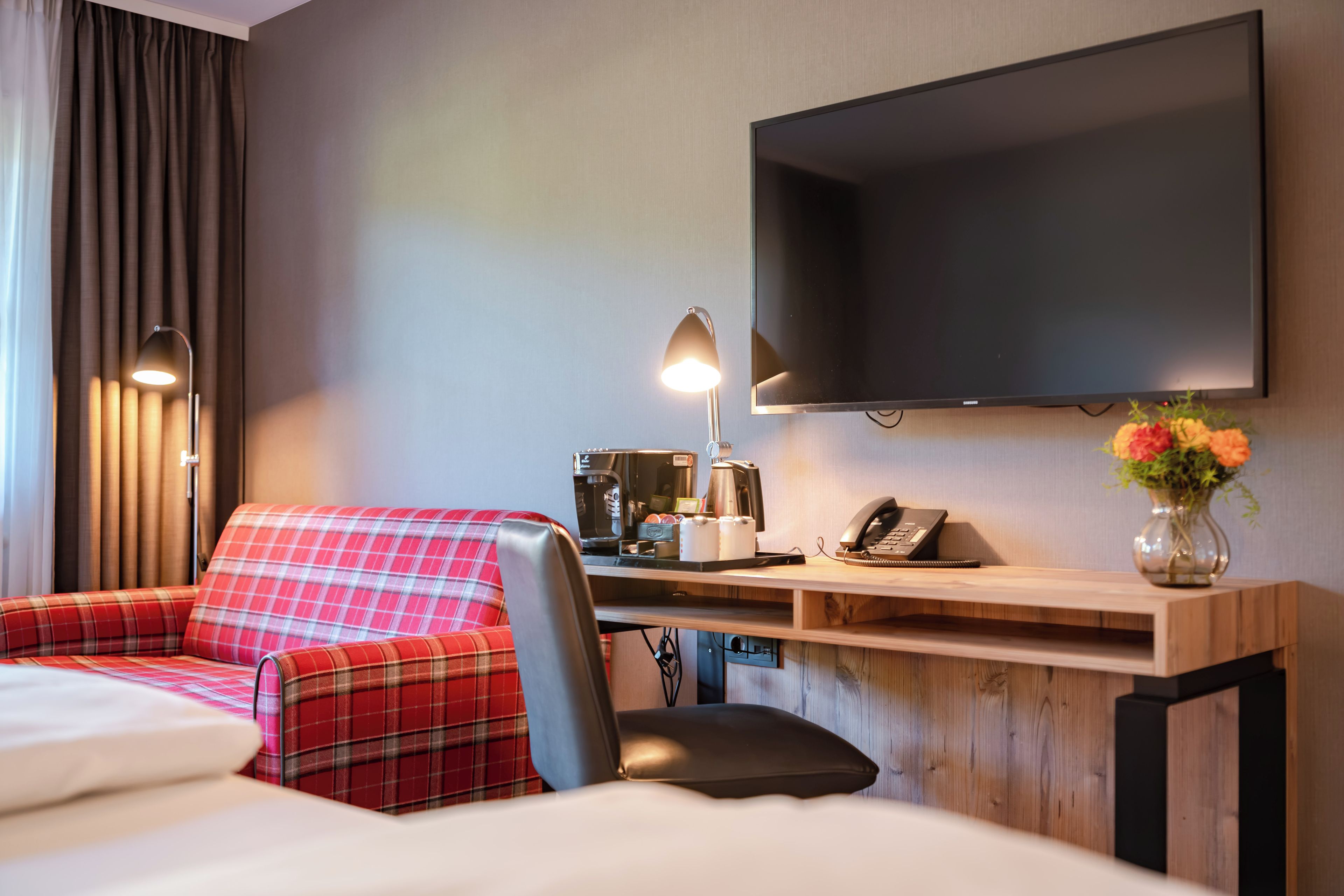 A hotel room with a bed, a red plaid sofa, a desk with a coffee maker, a telephone, and a flat-screen TV mounted on the wall.