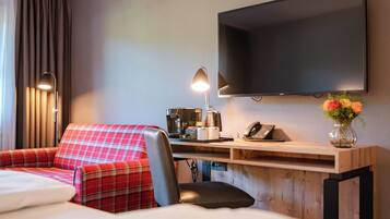 A hotel room with a bed, a red plaid sofa, a desk with a coffee maker, a telephone, and a flat-screen TV mounted on the wall.