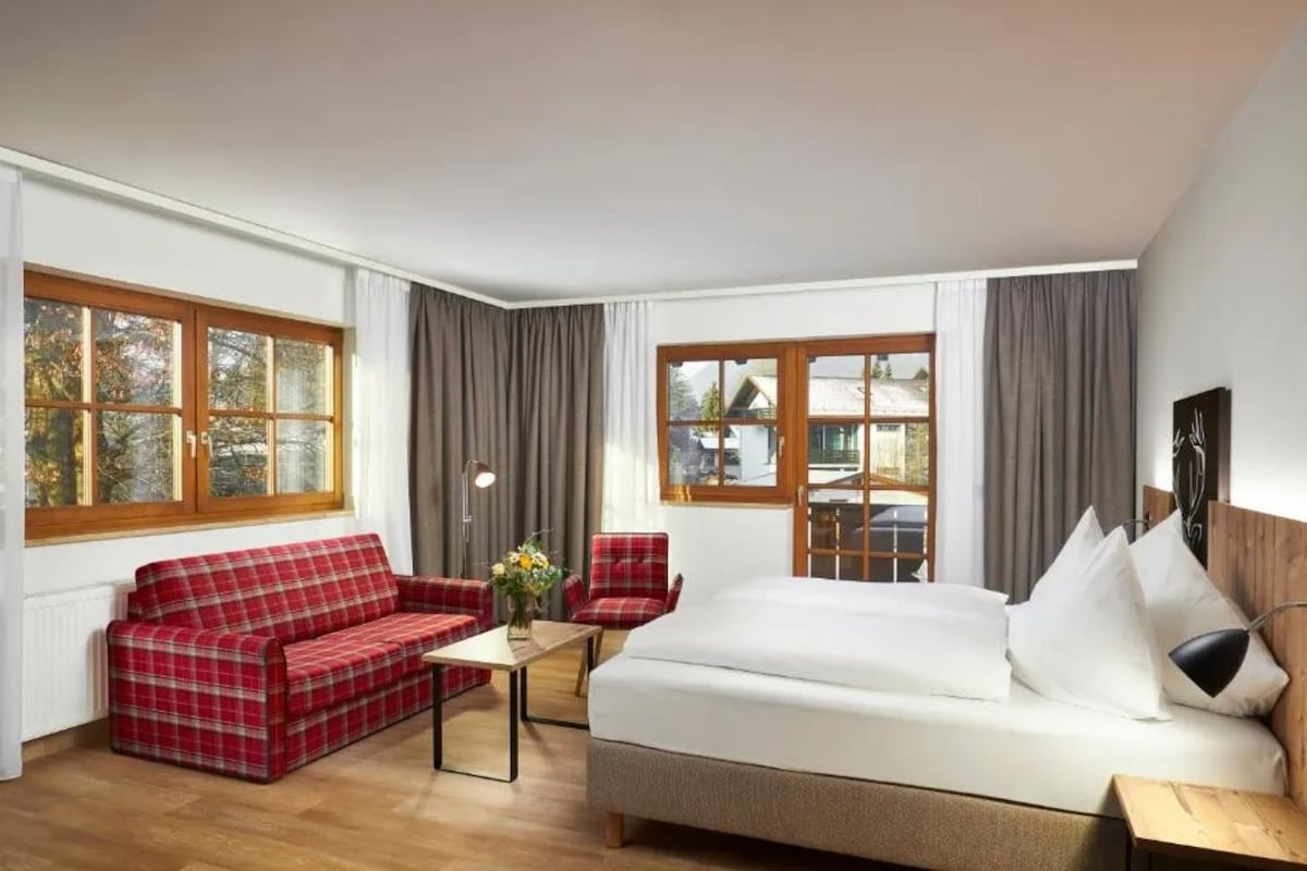 Hotellihuone, jossa on sänky, punavalkoruudullinen sohva, pieni pöytä ja kaksi tuolia.