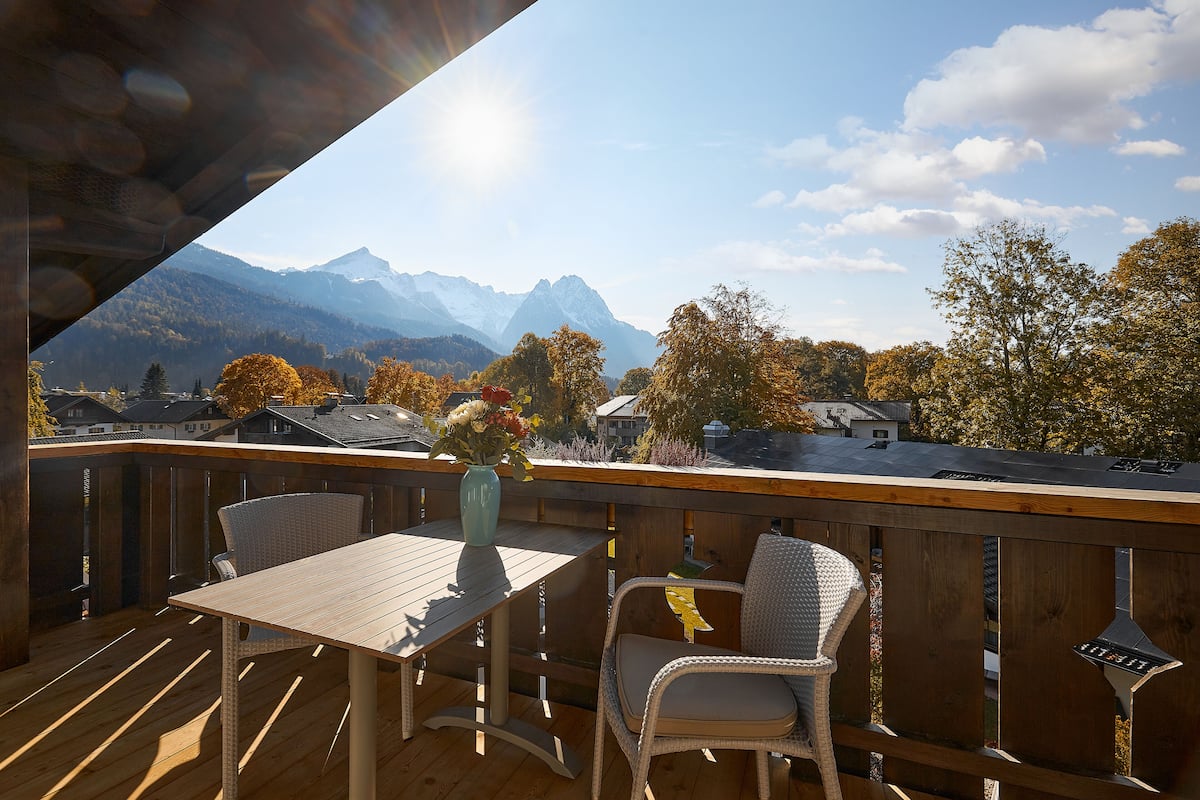 A wooden deck with a table and chairs, overlooking a mountainous landscape with autumn trees and a clear sky.